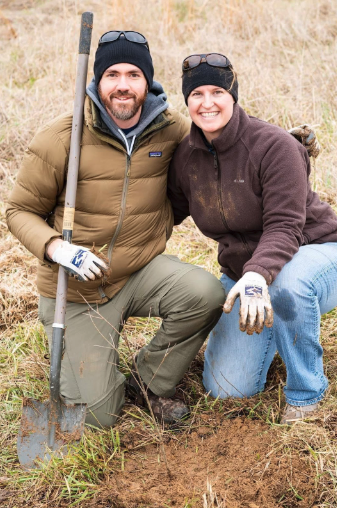 Two volunteers with Volunteer Odyssey crouching on the ground outdoors with a shovel, smiling at the camera, possibly taking a break from gardening or manual work, wearing casual work attire and gloves.